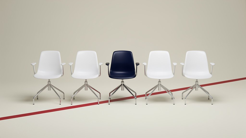 Five white office chairs in a row on a pale studio floor, the middle one navy blue, with a red stripe running diagonally across the floor.