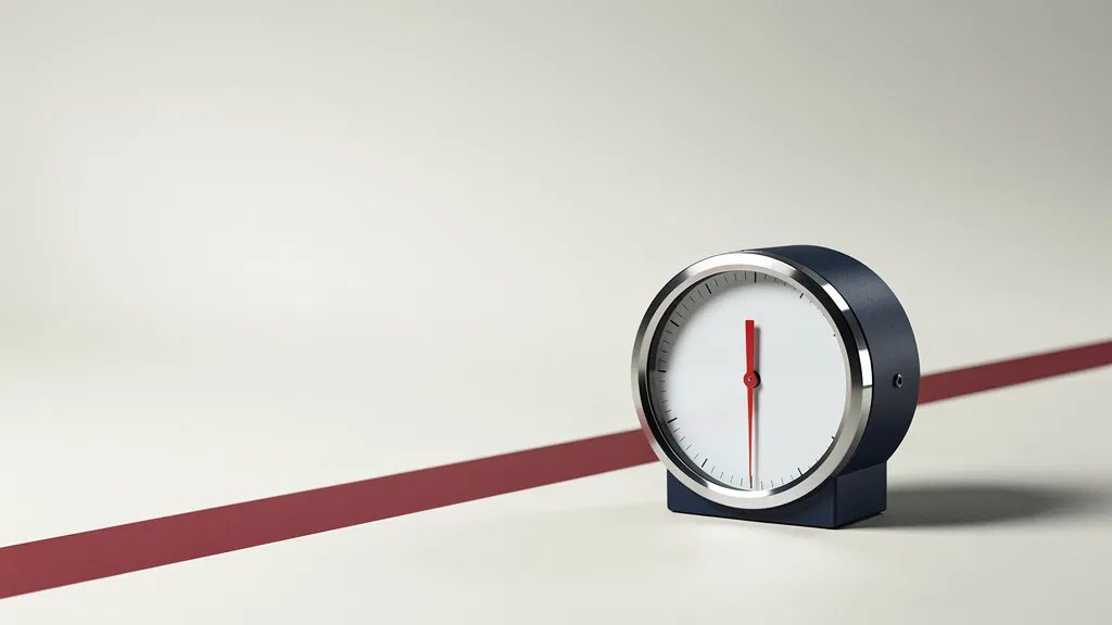 A navy analogue gauge with a white face and red needle on a pale floor, a red stripe behind it.